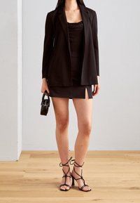 Czarny żakiet z otwartym przodem, nałożony na dopasowaną czarną sukienkę z bocznym rozcięciem. Dodatki to mała czarna torebka i sandały na obcasie.