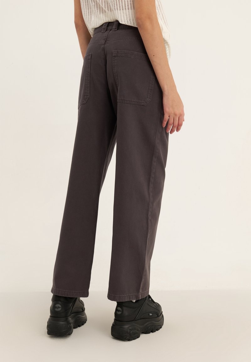 Pantalon gris à jambes larges avec poches arrière, fabriqué en tissu de coton durable. Porté avec des baskets noires épaisses.