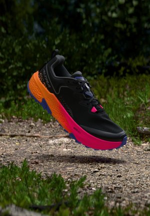 Zapatilla de correr negra con suela rosa brillante y naranja, flotando sobre un sendero de grava rodeado de vegetación verde.