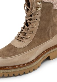 Stivale alla caviglia in suede e pelle marrone con chiusura lacci, occhiali metallici e suola spessa e texture. Fodera morbida visibile nella parte superiore.