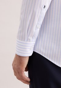 Biała koszula w paski z na przemian jasnoniebieskimi paskami; posiada mankiet na guziki oraz mały czarny haft na rękawie.