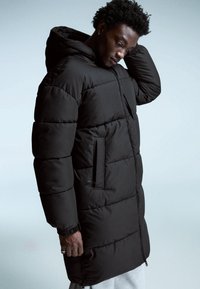 Cabană puffer neagră cu glugă, prevăzută cu o închidere cu fermoar și buzunare laterale. Materialul pare neted, cu un model matlasat pentru izolare.