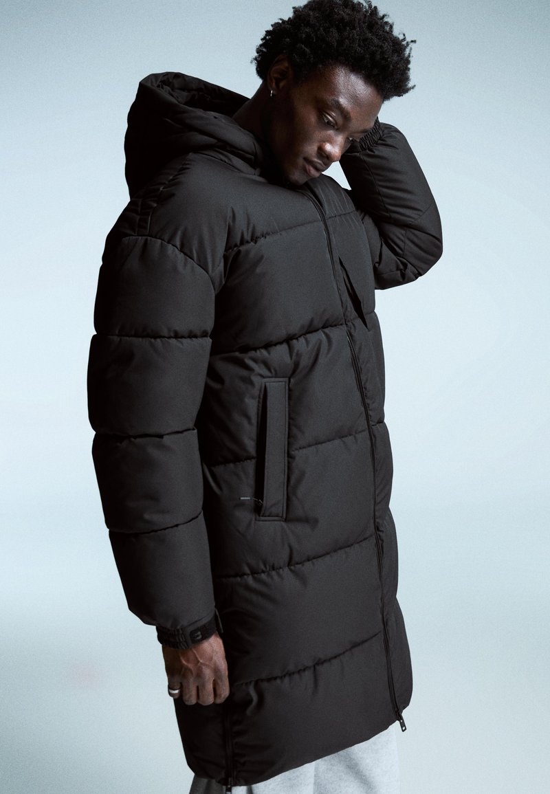 Cabană puffer neagră cu glugă, prevăzută cu o închidere cu fermoar și buzunare laterale. Materialul pare neted, cu un model matlasat pentru izolare.