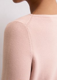 Jasnoróżowy sweter o fakturowanym dzianinowym wzorze i okrągłym dekolcie, przedstawiający zbliżenie szczegółów szwu na ramieniu i plecach.