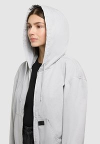 Jeune femme portant une veste à capuche gris clair par-dessus un t-shirt noir, tournée vers la gauche sur un fond gris uni.