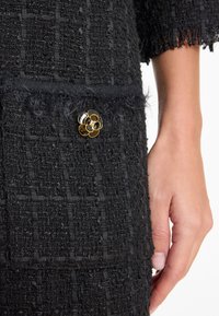 Zwart textiel met een geweven patroon. Bevat een bontafwerking en een gouden bloemvormige broche met zwarte en heldere accenten.