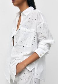 Chemise en coton blanc avec motif ajouré, fermeture boutonnée à l'avant, une poche poitrine unique et manches retroussées avec boutons décoratifs.