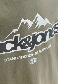 Olijfgroen katoenen T-shirt met een bergafbeelding in het wit, voorzien van vetgedrukte zwarte tekst met de woorden "jack & jones" en "STANDARD ISSUE SUPPLIES."