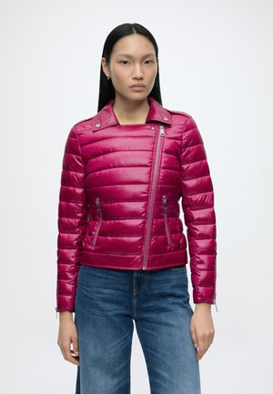 Vrouw met lang zwart haar, gekleed in een glanzend magenta doorgestikt jack met diagonale ritssluiting en blauwe spijkerbroek, staand tegen een witte achtergrond.