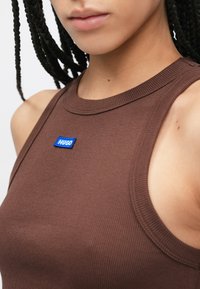 Καφέ αμάνικη μπλούζα με ριμπ υφή που διαθέτει ένα μπλε ορθογώνιο κεντητό τσόχινο "Hugo" στην περιοχή του στήθους. Ο σχεδιασμός περιλαμβάνει στρογγυλεμένο λαιμόκοψη και μανίκια χωρίς μανίκια.