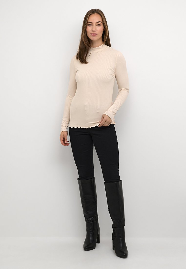 Langärmliges, geripptes Rollkragenoberteil in Beige mit gewelltem Saum, kombiniert mit schwarzen engen Hosen und knielangen schwarzen Stiefeln.