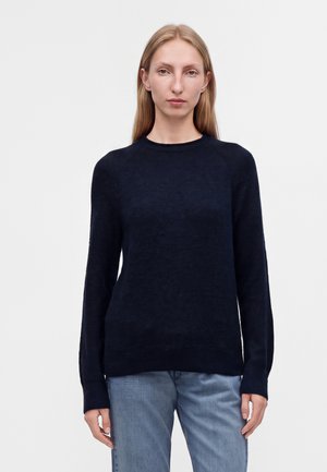 Femme aux longs cheveux blonds portant un pull bleu marine uni à manches longues et un jean bleu clair, debout sur un fond clair uni.