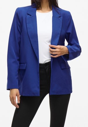 Femme portant un blazer bleu sur un top blanc et un pantalon noir, une main ajustant le blazer et des bijoux minimalistes aux doigts.