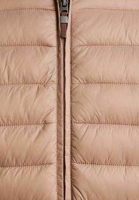 Kamizelka puffer w delikatnym beżowym kolorze, z poziomymi przeszyciami, mocnym zamkiem błyskawicznym i teksturowanym materiałem o gładkim wykończeniu.