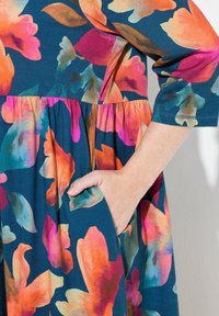 Květinové šaty s tmavě tyrkysovým základem a živými růžovými, oranžovými a zelenými vzory. Součástí je nařasený pas a boční kapsy.