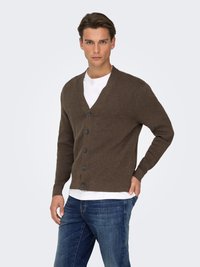 Cardigan marron tricoté avec un col en V, six boutons et des manches longues, porté sur un T-shirt blanc, associé à un jean bleu.