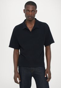 Polo shirt închis la culoare, cu mânecă scurtă, de nuanță bleumarin închis, cu un model texturat, guler pliat și o croială lejeră, asortat cu blugi din denim închis.