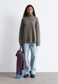 Übergroßer grauer Pullover mit gerippten Bündchen, hellblaue Jeans, schwarze klobige Schuhe und ein lila-blauer Fransen-Schal, der in einer Tasche gehalten wird.