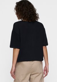 Top in maglia nera a maniche corte con vestibilità ampia e scollo a girocollo. Presenta una cucitura centrale sul retro. Indossato con pantaloni beige.