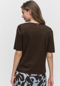 T-shirt marron à manches courtes avec un col rond et un petit accent circulaire à l'arrière, fabriqué en tissu de coton lisse.