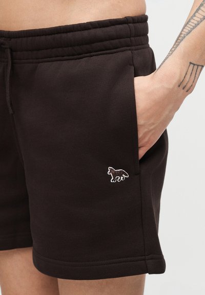 Κοντινό πλάνο ενός ατόμου που φοράει σκουρόχρωμα σορτς με ένα μικρό κεντημένο λογότυπο αλεπούς κοντά στην τσέπη και τατουάζ στον πήχη.