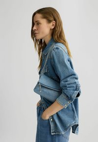 Borsa a spalla in denim con una finitura blu texturizzata, dotata di tracolla in catena argentata e chiusura a zip, tenuta contro un giubbotto di denim.