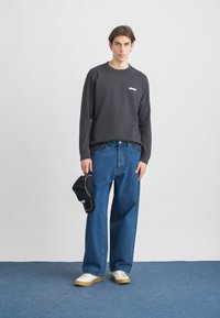 Sweatshirt de mangas longas cinza escuro com logótipo branco, combinado com jeans azuis largos e sapatilhas brancas. Segura uma cap preto.