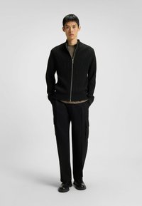 Giovane uomo in piedi con le mani in tasca, che indossa un maglione zip-up a costine nero, pantaloni cargo neri e scarpe nere su uno sfondo grigio chiaro.