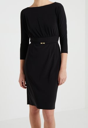 Kobieta ubrana w dopasowaną czarną sukienkę do kolan z rękawami do łokcia i cienkim czarnym paskiem ze złotą klamrą w talii.