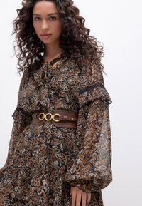 Robe fleurie marron avec manches à volants, équipée d'une ceinture marron avec accessoires dorés et un détail noué au niveau de l'encolure.