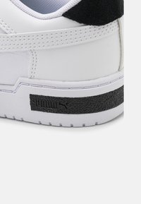 Vit Puma sneakerklack med svart texturerad logotre överdrag och sydda detaljer på en vit sula.