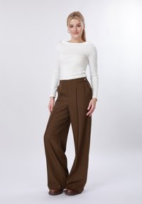 Jeune femme debout, portant un haut blanc à manches longues, un pantalon marron taille haute à jambes larges, et des chaussures marron, sur un fond clair uni.