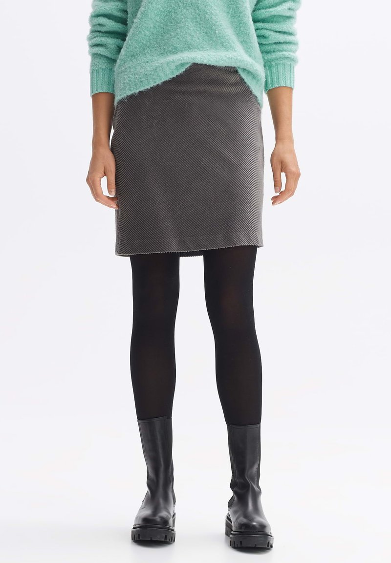 Grauer Cordrock mit schrägem Saum, kombiniert mit schwarzen blickdichten Strumpfhosen und schwarzen Knöchelstiefeln. Strukturiertes Material sichtbar.