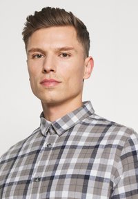 Grå og hvit rutete skjorte med krage, laget av teksturert stoff og med grå knapper, vist på en mannlig modell.