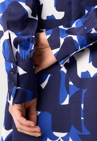 Mains croisées dans le dos, portant un vêtement à manches longues à motif abstrait bleu, blanc et noir avec des poignets boutonnés.