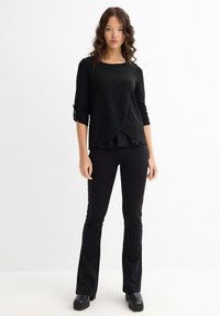 Top negro de textura envolvente con mangas largas y detalles fruncidos, combinado con pantalones negros ajustados de corte acampanado y zapatos negros gruesos.