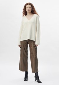 Übergroßer cremfarbener Pullover mit V-Ausschnitt, kombiniert mit braunen Leder-Cargohosen, die über Seitentaschen und gestickte Details verfügen. Schwarze Stiefeletten mit Absatz.