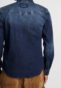 Camicia in denim scuro con una sottile texture usurata, dotata di un design a yoke e polsini con bottoni. Orlo curvo con spacco laterale per una maggiore facilità.