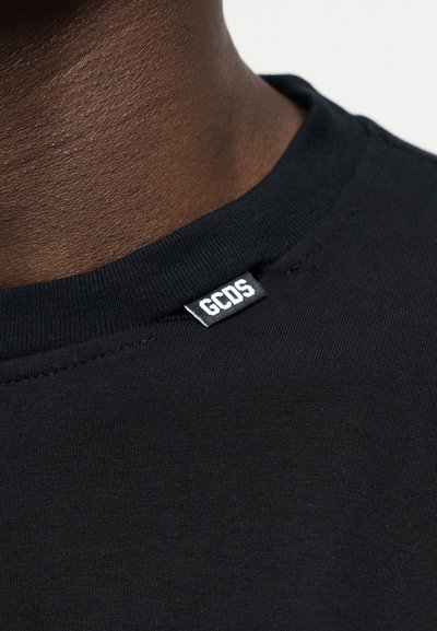 Κοντινό πλάνο του λαιμού ενός ατόμου με σκούρο δέρμα, που φοράει μαύρη μπλούζα με στρογγυλή λαιμόκοψη, με μια ορατή λευκή ετικέτα "GCDS" στον γιακά.