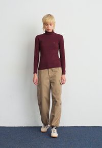 Persona con capelli biondi corti che indossa un maglione a collo alto bordeaux, pantaloni cargo beige e sneaker bianche in piedi contro un muro bianco.