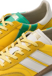 Paire de chaussures de sport jaunes et blanches avec des accents en daim, des lacets jaunes et une doublure intérieure verte, marquées "ANIMAL" sur la languette.