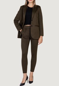 Blazer de espiga en color oliva con un solo botón, combinado con pantalones ajustados. Top corto de canalé negro debajo. Zapatos de tacón negros completan el look.