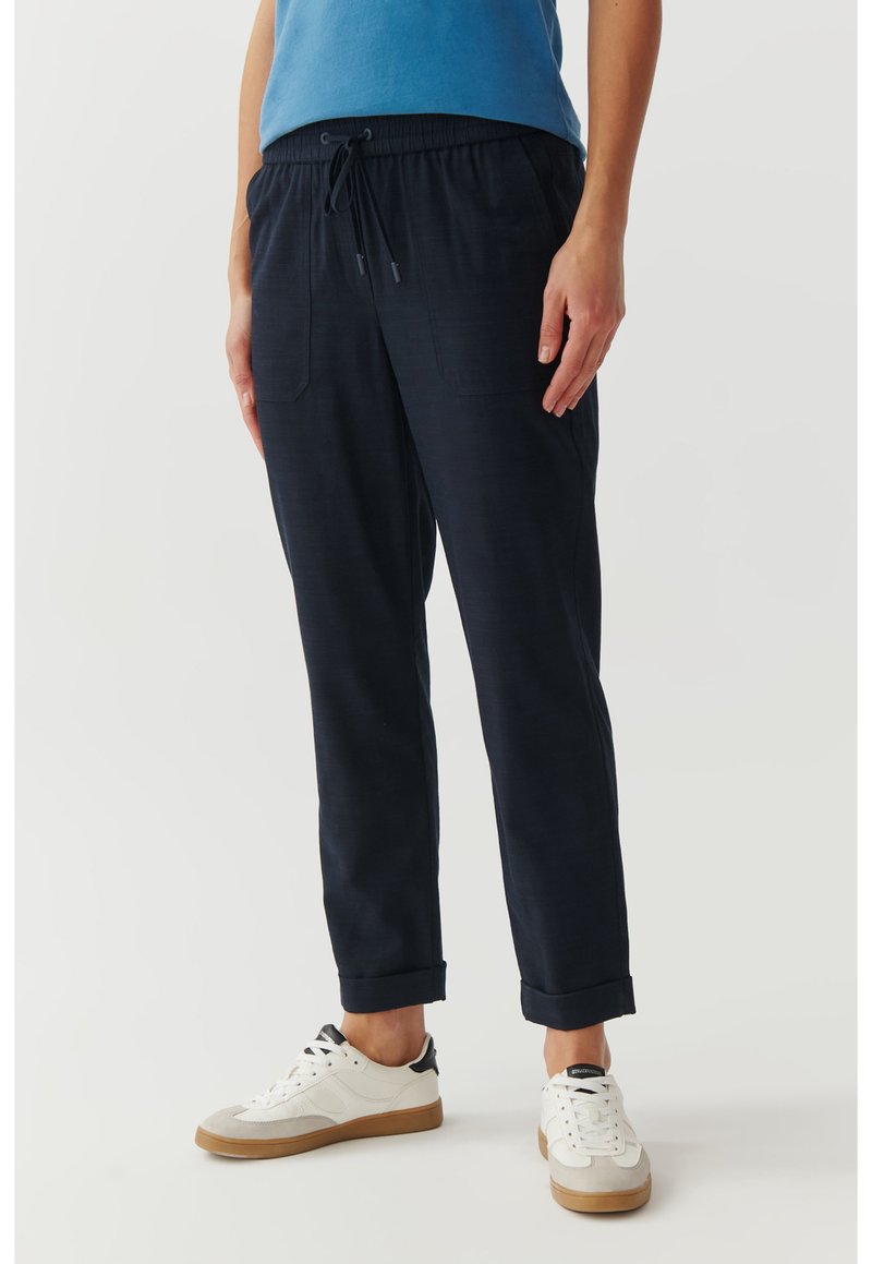 Marineblaue Hose aus leichtem Stoff mit elastischem Bund, zwei Vordertaschen und umgeschlagenen Säumen. Trägt weiße und graue Sneaker.