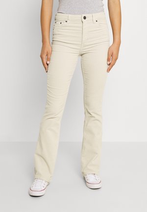 Woman wearing light beige straight-leg pants and white sneakers, standing against a plain white background.