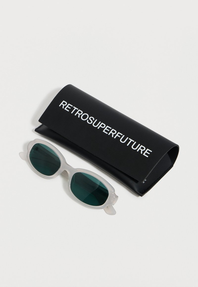 Gafas de sol ovaladas con montura blanca y lentes verde oscuro junto a un estuche negro con la etiqueta RETROSUPERFUTURE sobre una superficie blanca.