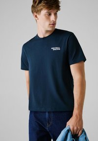 Marineblauwe katoenen T-shirt met een witte "RED BULL RACING" logo op de linkerkant van de borst, ronde hals en korte mouwen, gecombineerd met donkere spijkerbroek.