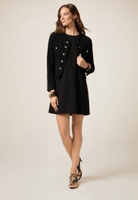 Schwarze kurze Jacke mit goldenen Knöpfen, kombiniert mit einem schwarzen Kleid. Modell trägt schwarze High Heels und ein Armband. Glatte Texturen sind sichtbar.