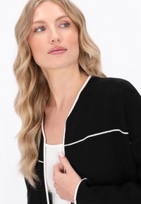 Schwarze Jacke mit weißem Piping entlang der Vorderseite. Der Stoff wirkt weich, mit einer lässigen Passform und offenem Design. Das Model hat lange, wellige Haare.