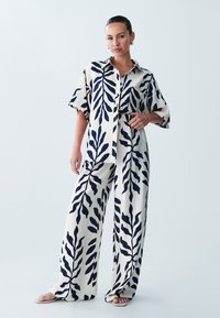 Dvoudílný outfit skládající se z košile na knoflíky s krátkým rukávem a širokými kalhotami. Krémový základ s námořnickým listovým vzorem, vyrobený z lehkého materiálu.
