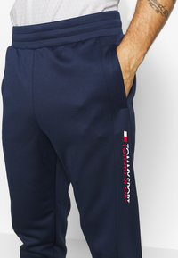 Pantaloni sportivi blu navy con una texture liscia, caratterizzati da un logo verticale di marca rosso e bianco sul lato sinistro e una vita elastica.